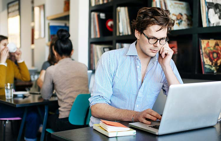 Man sitting at a table in an internet cafe on his personal laptop using the internet on public a wifi network.